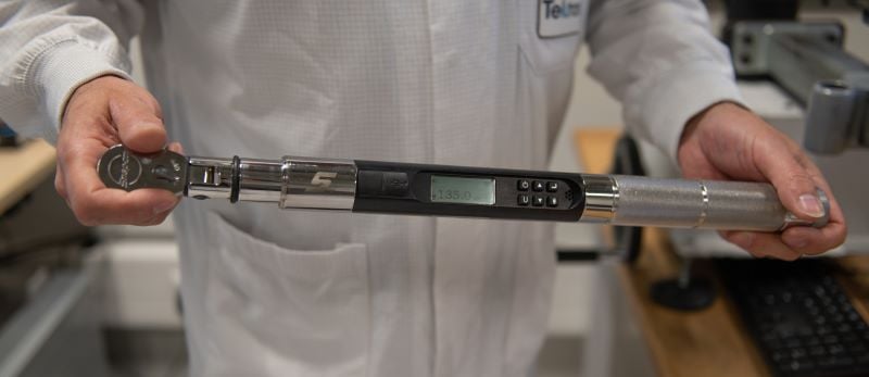 A calibration technician in a white lab coat holds a digital torque wrench horizontally, with the display screen showing 
