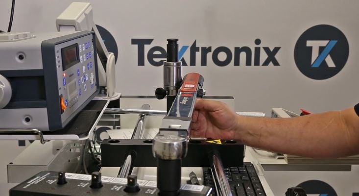 A calibration technician operates torque wrench calibration equipment, featuring a digital display unit with buttons and indicators connected to a mechanical fixture that securely holds the torque wrench. The Tektronix logo is visible in the background.
