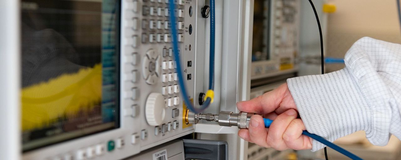 Close-up of a technician's hand inserting a connector into an oscilloscope for calibration. The oscilloscope displays a waveform on its screen, with cables and connectors visible around the device.