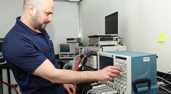 A technician operating an oscilloscope in a laboratory setting with various electronic equipment on shelves