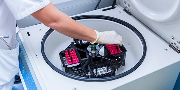 A laboratory technician’s gloved hand is placing test tube racks filled with red-capped tubes into a centrifuge machine